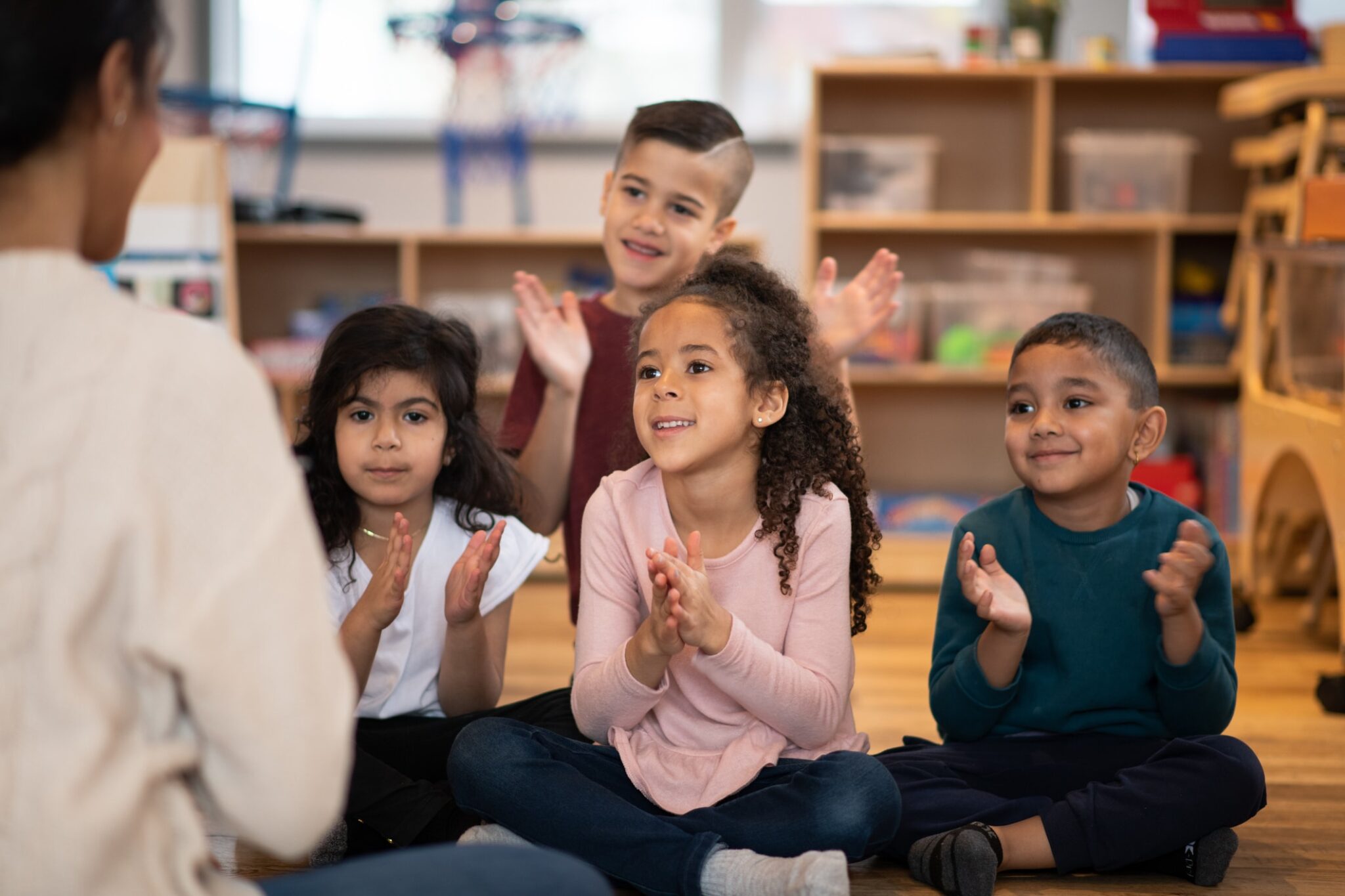 A young female teacher of Indian decent, leads a small group of multi-ethnic daycare children in a series of songs. The children are each dressed casually and sitting cross-legged in front of their teacher as they, smile, clap and sing along. The focus is on a sweet Indian boy in front.