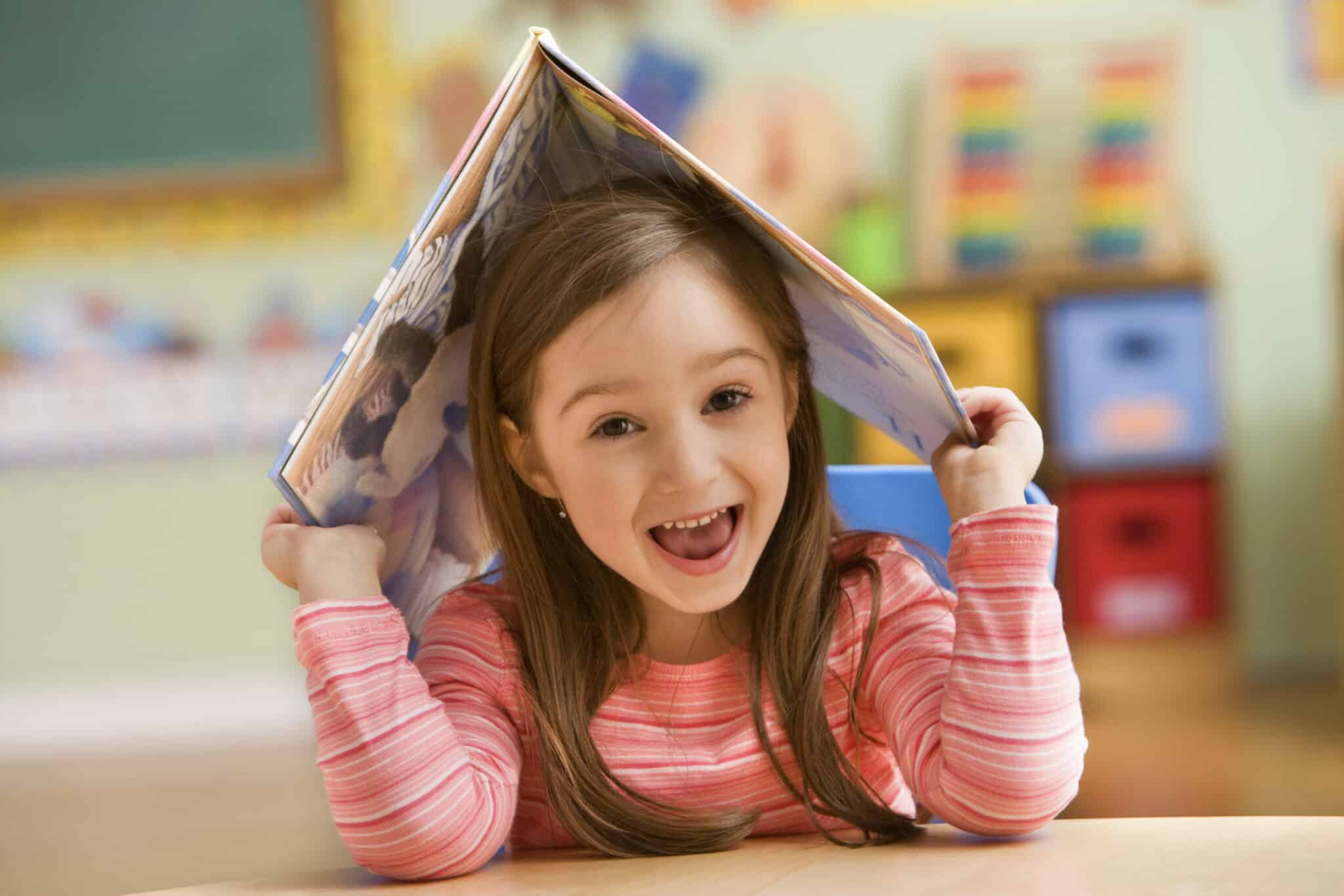 Girl (4-5) holding open book on head and smiling with open mouth