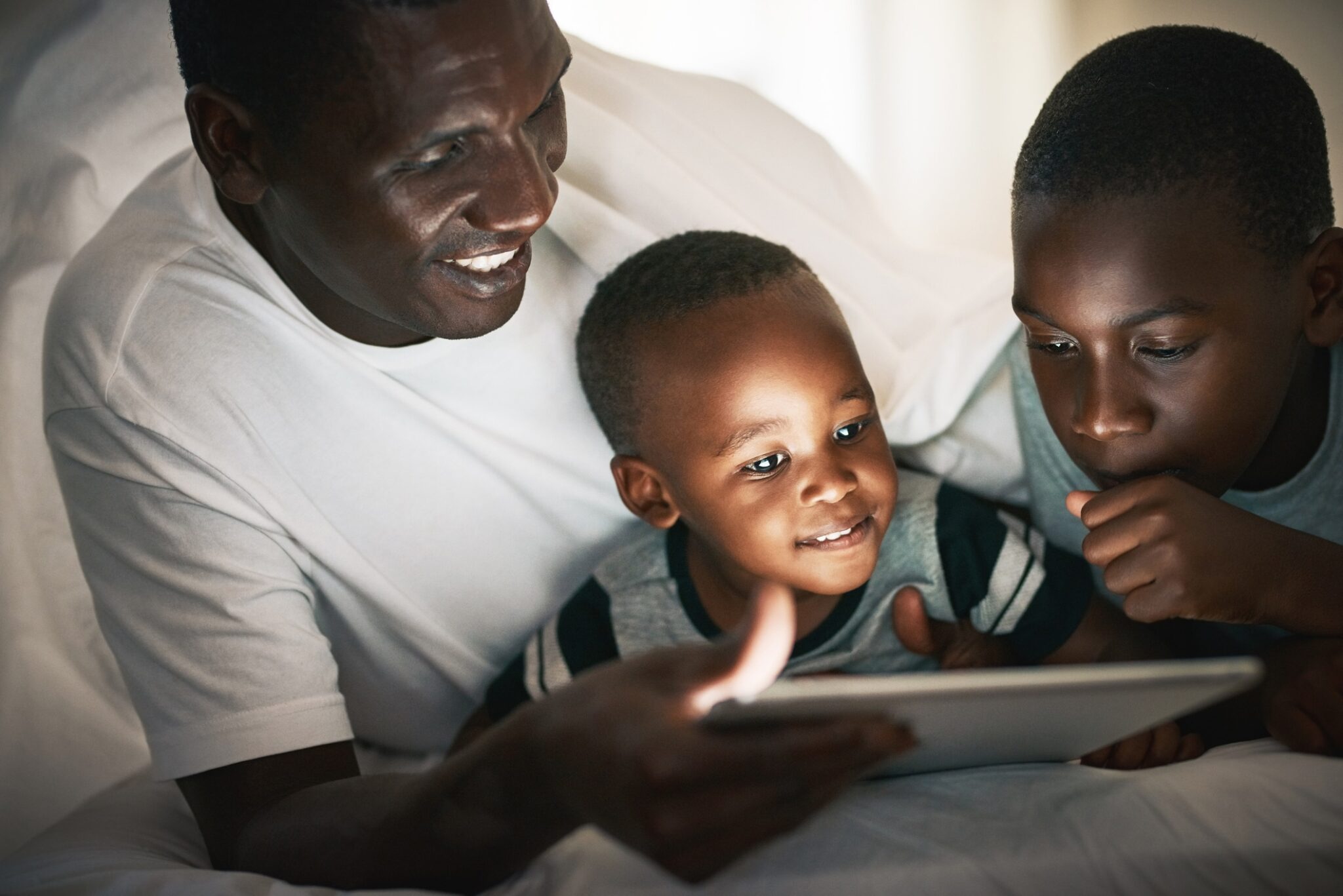 young boy using a digital tablet with his father and brother at night in bed